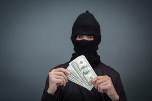 A person wearing a black ski mask holding a fan of U.S. dollar bills, representing scams or fraudulent activity.