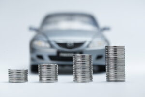 Stacks of coins increasing in height in front of a blurred car, representing rising vehicle costs or savings for a car purchase.
